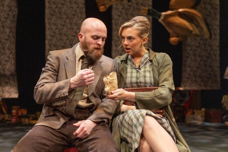 Blonde woman site next to a bald man with a beard in a theatre production of Happy Jack