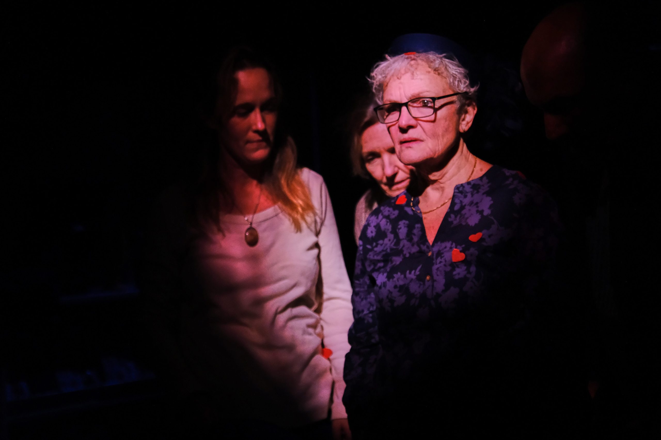An older woman looks off into the distance with two other women in the shadows behind her