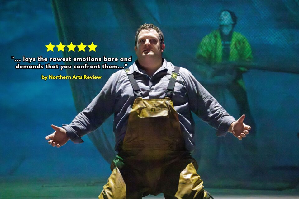 Fisherman in waders stands on the stage with his arms wide open from opera Peter Grimes at Leeds Grand by Opera North